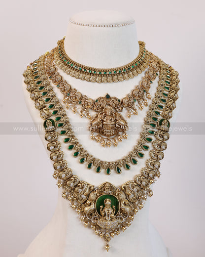 An ornate multi-layered gold-toned traditional necklace set displayed on a white mannequin bust. The jewelry features green stone accents, detailed gold filigree, and a central deity motif with a green oval background.